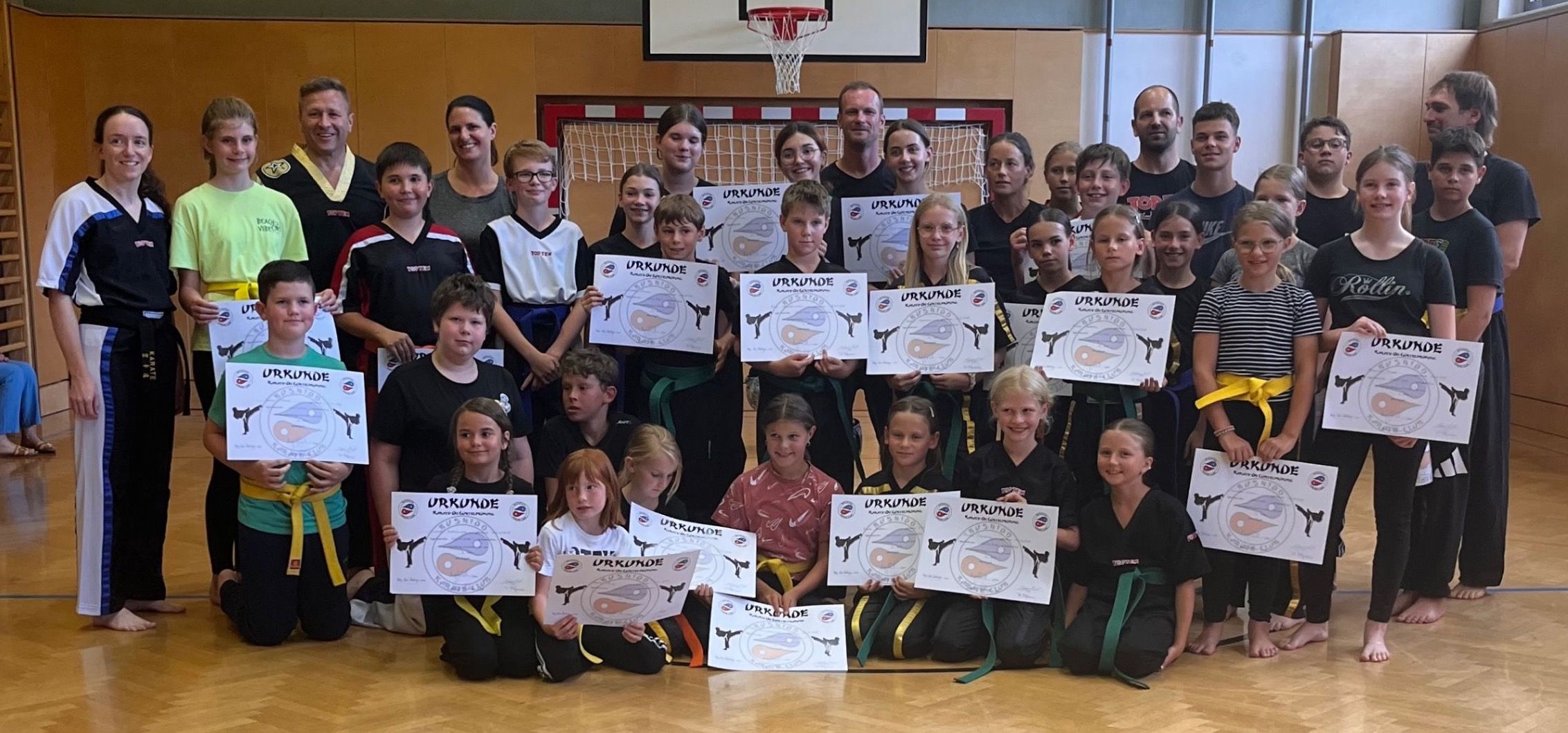Eine Gruppe junger Kampfkünstler in schwarzen und weißen Uniformen posiert mit Urkunden in einem Fitnessstudio.