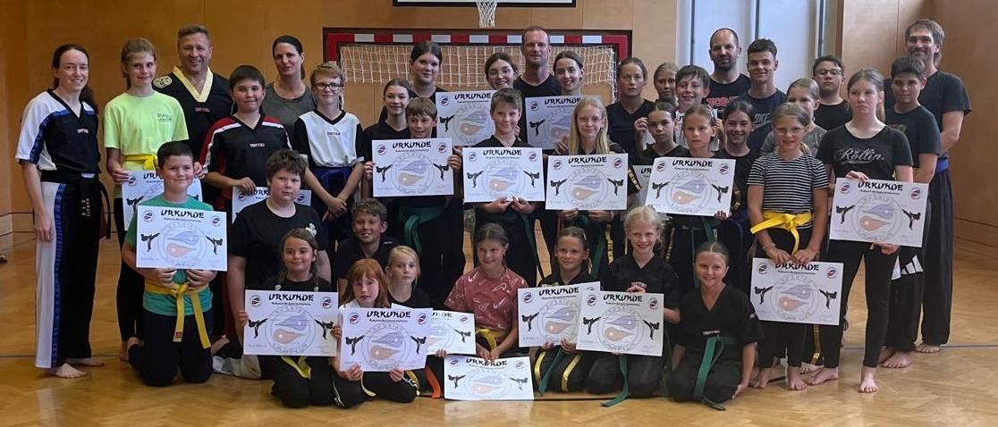 Eine Gruppe von Kindern und Erwachsenen, die Kampfsportuniformen tragen, posiert für ein Foto und hält Urkunden in der Hand. Sie befinden sich in einem Raum mit Holzboden und einem Basketballkorb.