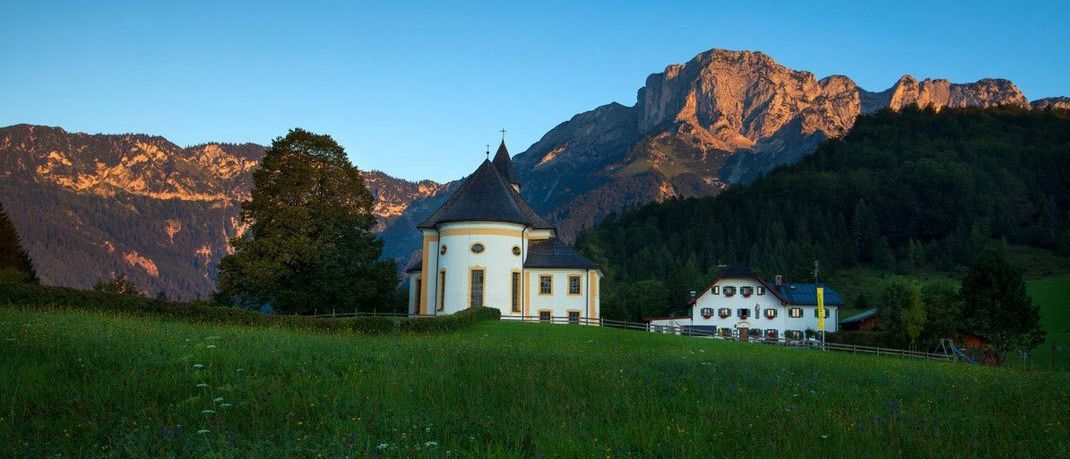 Eine Kapelle steht auf einem grasbewachsenen Hügel unter einem klaren blauen Himmel, mit Bergen im Hintergrund.