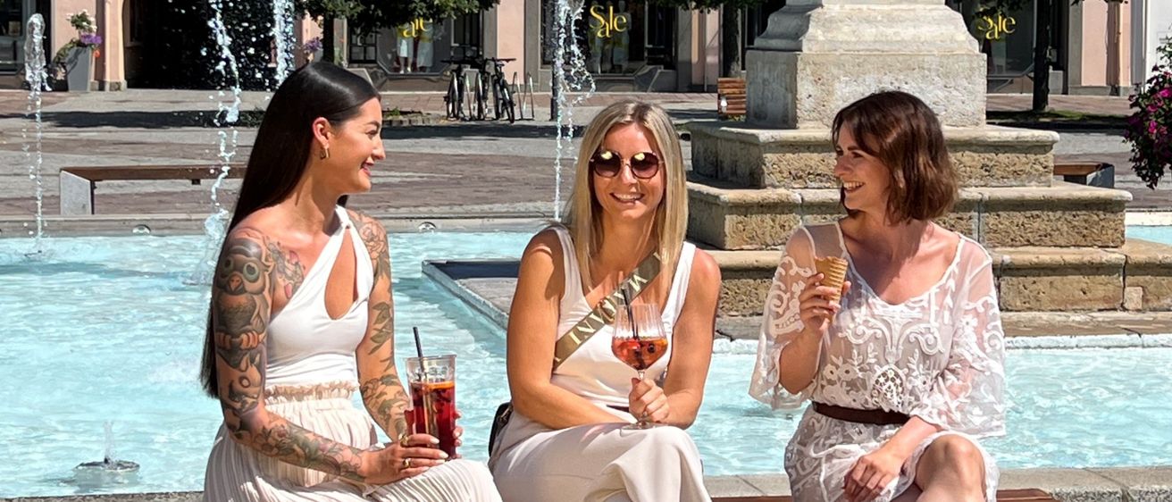 Drei Frauen in weißen Kleidern sitzen auf einer Bank neben einem Brunnen. Eine hält ein Weinglas, eine andere einen Cocktail und die dritte hat Einkaufstaschen. Hinter ihnen steht ein Gebäude mit einem roten Dach und einem großen Steinmonument.