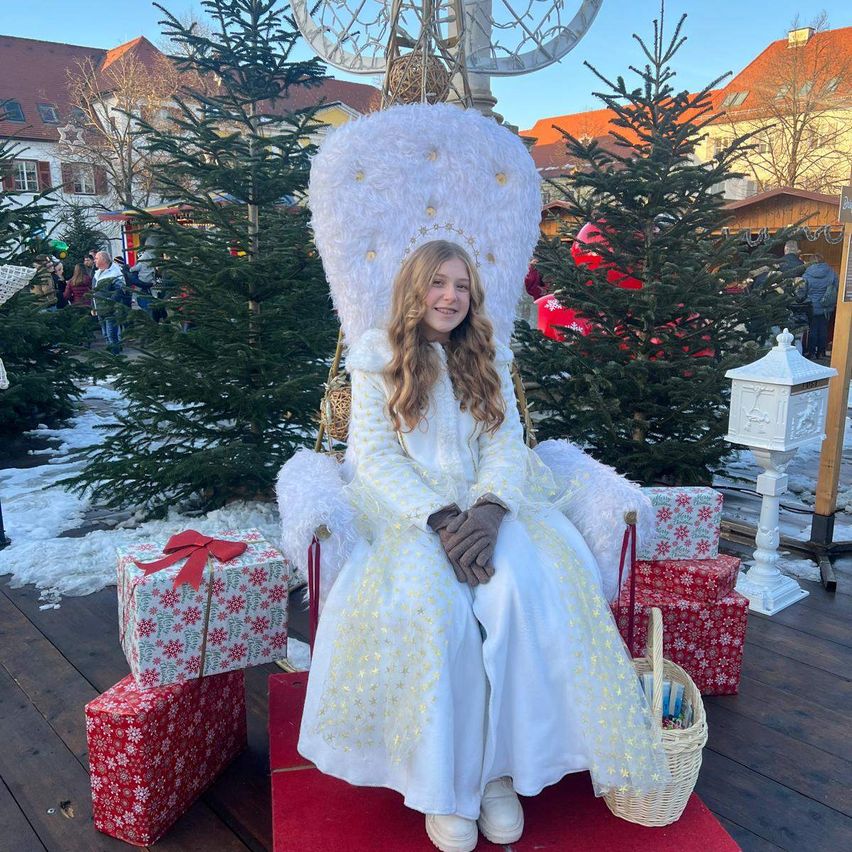 Eines Mädchen in einem weißen Kleid sitzt auf einem Thron, der mit weißem Pelz und goldenen Sternen verziert ist, umgeben von Weihnachtsgeschenken. Hinter ihr stehen Weihnachtsbäume und Gebäude im Hintergrund.