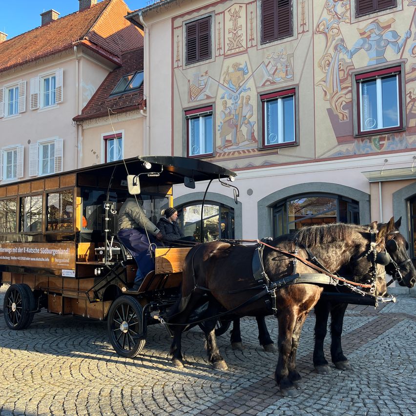Eine Pferdekutsche mit Passagieren steht auf einer Kopfsteinpflasterstraße vor einem Gebäude mit Wandgemälden.