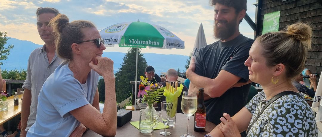 Eine Gruppe von Menschen steht um einen Tisch auf einer Terrasse und lächelt. Ein Regenschirm steht hinter ihnen und eine Frau hält ein Weinglas. Eine andere Frau hat ihr Kinn auf der Hand.