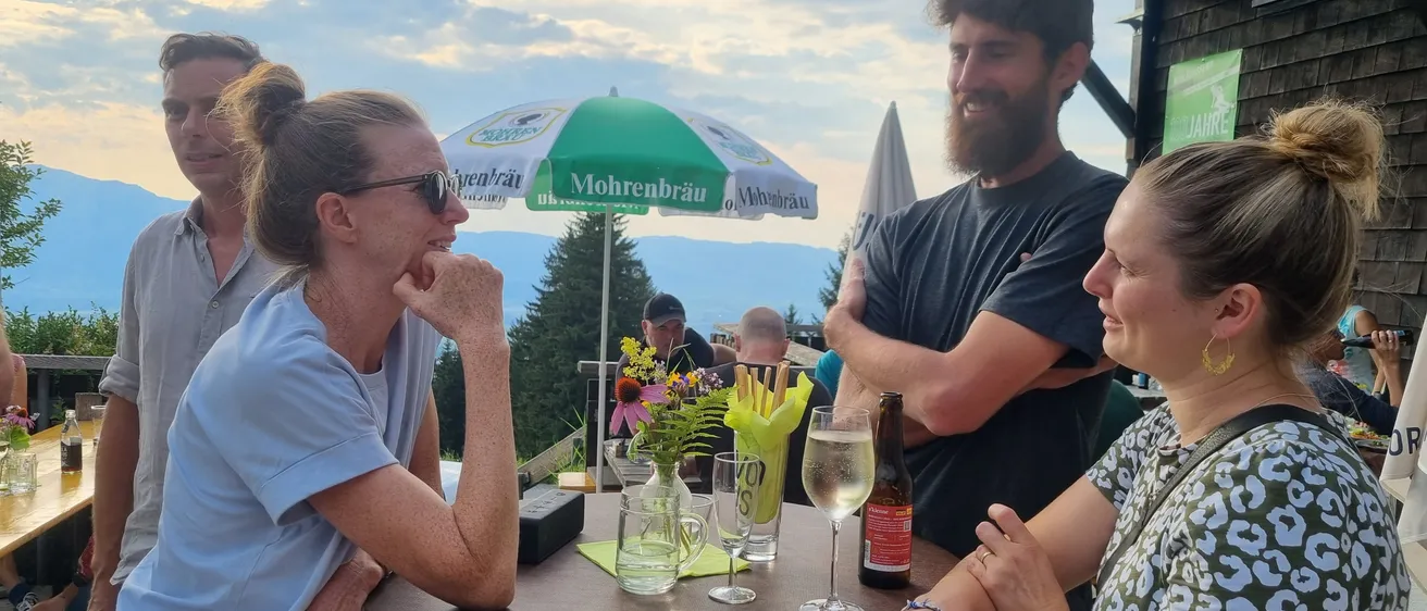 Eine Gruppe von Menschen steht um einen Tisch auf einer Terrasse und lächelt. Ein Regenschirm steht hinter ihnen und eine Frau hält ein Weinglas. Eine andere Frau hat ihr Kinn auf der Hand.