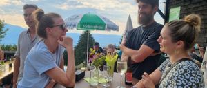 Eine Gruppe von Menschen steht um einen Tisch auf einer Terrasse und lächelt. Ein Regenschirm steht hinter ihnen und eine Frau hält ein Weinglas. Eine andere Frau hat ihr Kinn auf der Hand.