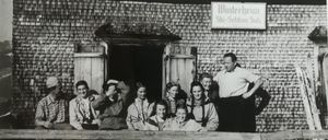 Ein Schwarz-Weiß-Foto zeigt eine Gruppe von Menschen, die vor einem Gebäude namens 'Winterheim' stehen und sitzen. Die Menschen sind in Winterkleidung gekleidet, einige mit Mützen. Ein Mann steht vor der Gruppe mit den Händen auf den Hüften.