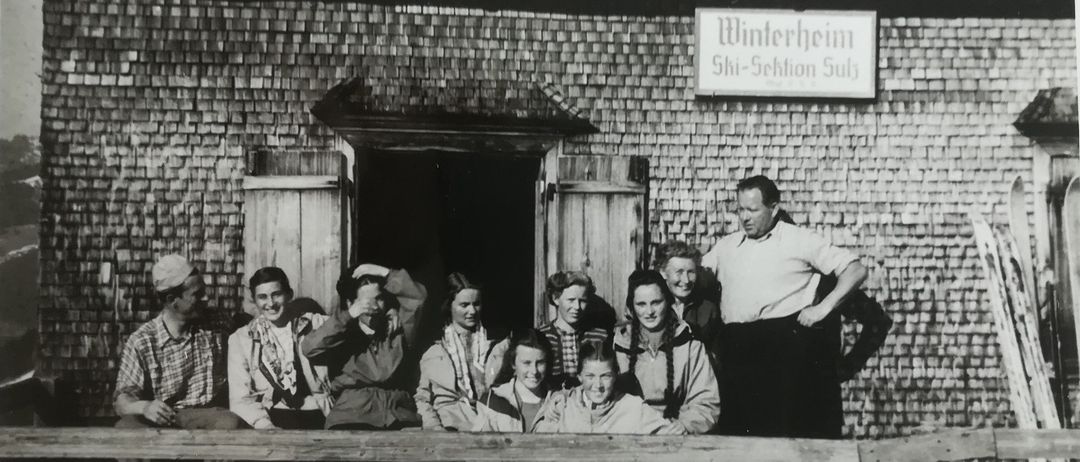 Ein Schwarz-Weiß-Foto zeigt eine Gruppe von Menschen, die vor einem Gebäude namens 'Winterheim' stehen und sitzen. Die Menschen sind in Winterkleidung gekleidet, einige mit Mützen. Ein Mann steht vor der Gruppe mit den Händen auf den Hüften.