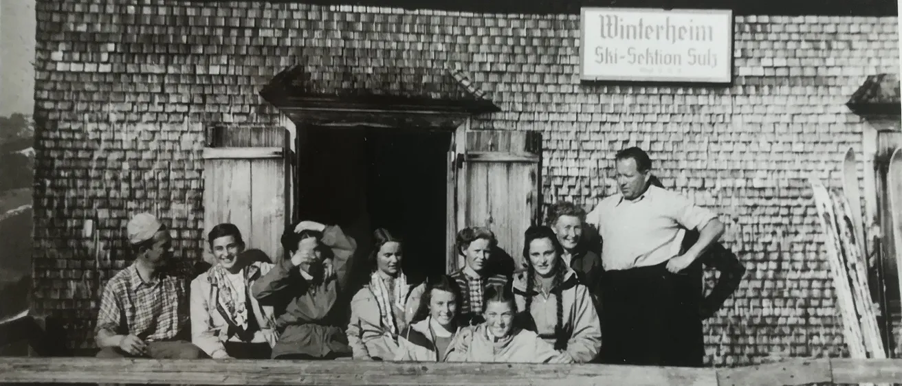 Ein Schwarz-Weiß-Foto zeigt eine Gruppe von Menschen, die vor einem Gebäude namens 'Winterheim' stehen und sitzen. Die Menschen sind in Winterkleidung gekleidet, einige mit Mützen. Ein Mann steht vor der Gruppe mit den Händen auf den Hüften.