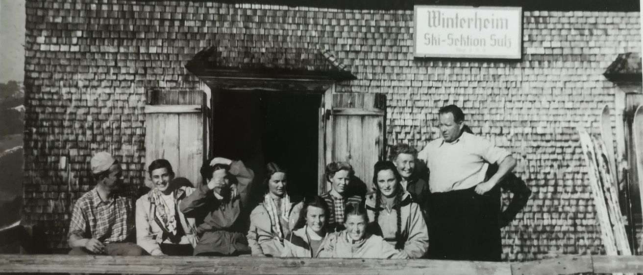 Ein Schwarz-Weiß-Foto zeigt eine Gruppe von Menschen, die vor einem Gebäude namens 'Winterheim' stehen und sitzen. Die Menschen sind in Winterkleidung gekleidet, einige mit Mützen. Ein Mann steht vor der Gruppe mit den Händen auf den Hüften.