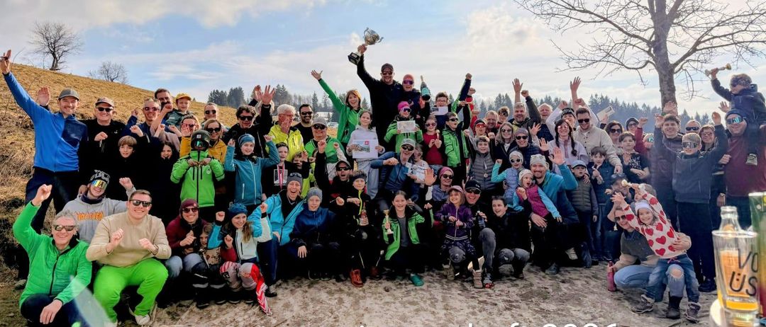 Eine große Gruppe von Menschen in Winterkleidung feiert im Freien. Ein Mann in der Mitte hält eine Trophäe, und andere halten Urkunden. Die Gruppe lächelt und posiert für ein Foto. Der Text lautet 'Vereinsmeisterschaft 2026'.