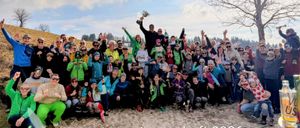 Eine große Gruppe von Menschen in Winterkleidung feiert im Freien. Ein Mann in der Mitte hält eine Trophäe, und andere halten Urkunden. Die Gruppe lächelt und posiert für ein Foto. Der Text lautet 'Vereinsmeisterschaft 2026'.