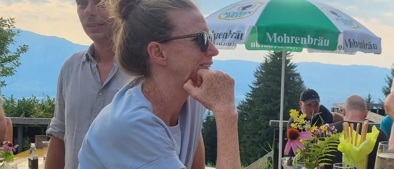 Eine Frau sitzt an einem Tisch mit einem Mann, lächelt und stützt ihr Kinn auf ihre Hand. Sie sind von anderen Menschen umgeben, einschließlich eines Mannes mit Hut und eines Mannes, der ein Getränk in der Hand hält. Es gibt einen Tisch mit einer Flasche, einem Blumenvase und einem Weinglas. In der Ferne gibt es eine Aussicht auf Berge.