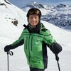 Ein Mann steht auf einem verschneiten Berg mit Bergen im Hintergrund. Er trägt eine grüne Skijacke, eine Brille und hält Skistöcke.