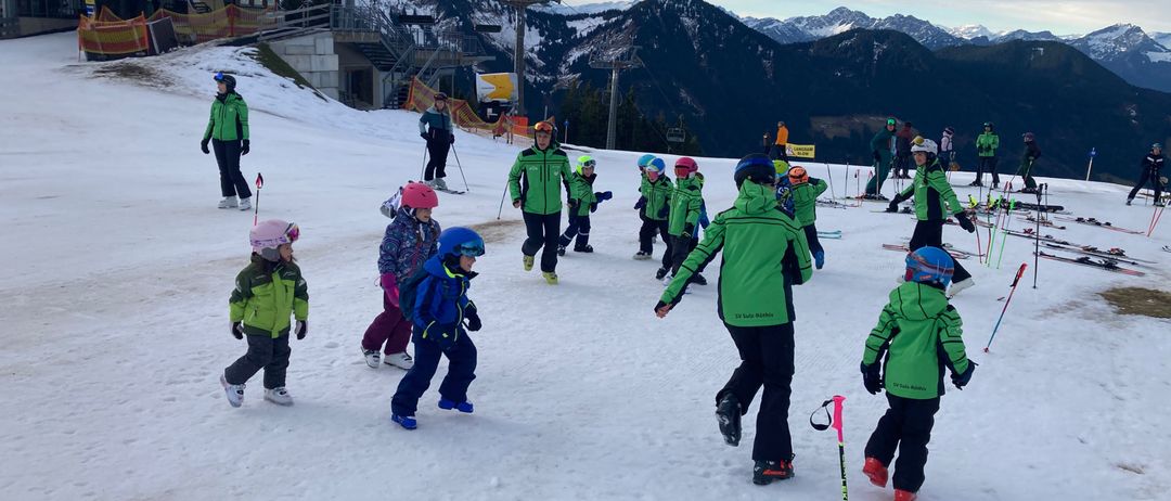 Eine Gruppe von Kindern in grüner Skiausrüstung lernt auf einer verschneiten Piste Skifahren, mit Bergen im Hintergrund.