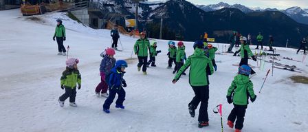 Eine Gruppe von Kindern in grüner Skiausrüstung lernt auf einer verschneiten Piste Skifahren, mit Bergen im Hintergrund.