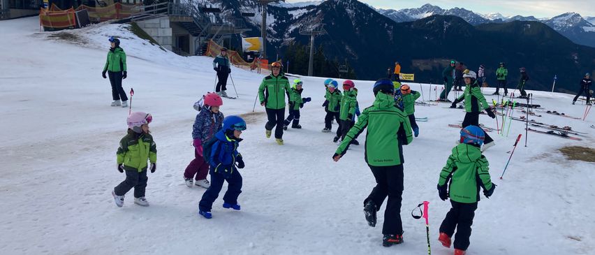 Eine Gruppe von Kindern in grüner Skiausrüstung lernt auf einer verschneiten Piste Skifahren, mit Bergen im Hintergrund.