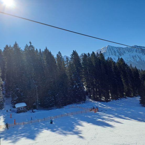 Eine verschneite Skipiste mit einer kleinen Hütte und mehreren Skifahrern unter einem klaren blauen Himmel mit Bergen im Hintergrund.