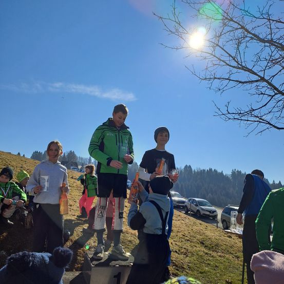 Unter einem klaren blauen Himmel steht ein junger Athlet auf einem Podest und hält eine Trophäe. Hinter ihm hält ein weiterer junger Athlet ein Glas. Zuschauer, darunter Kinder, versammeln sich auf dem Grashang, Autos geparkt in der Nähe.