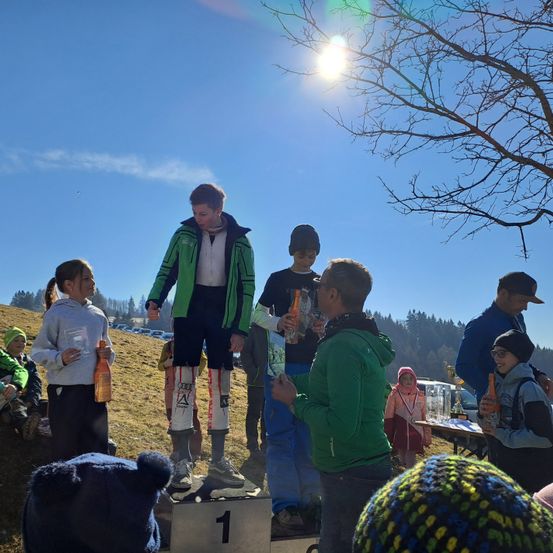 Eine Gruppe von Menschen versammelt sich an einem sonnigen Tag für einen Wettbewerb. Ein Mann steht auf der Spitze eines Podests mit der Nummer 1. Andere stehen um ihn herum, einige halten Trophäen und Getränke.