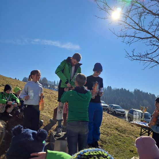Eine Gruppe von Menschen versammelt sich auf einem Grashügel unter einem klaren blauen Himmel, möglicherweise für einen Wettbewerb. Ein Junge steht auf einem Podium, während ihm ein anderer Junge einen Pokal überreicht. Die anderen beobachten. Im Hintergrund sind Autos am Straßenrand geparkt.