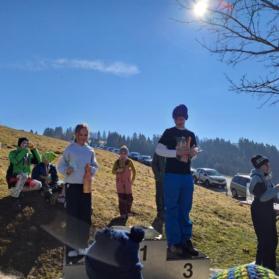 Mehrere Menschen stehen auf einem grasbewachsenen Hügel unter einem hellen Himmel. Drei Menschen stehen auf Podests, einer von ihnen hält eine Trophäe. Andere sitzen hinter ihnen, und Autos sind in der Nähe geparkt.