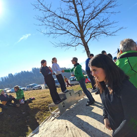 Zwei Mädchen stehen auf einem Podest und halten Trophäen. Ein drittes Mädchen steht neben ihnen. Dahinter beobachtet eine Menge Leute. Ein Baum und Berge sind im Hintergrund.