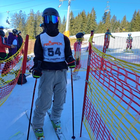 Ein Skifahrer mit der Nummer 54 steht in der Nähe einer Netzbarriere auf einer Skipiste, trägt einen Helm und eine Schutzbrille. Mehrere andere Skifahrer sind im Hintergrund zu sehen.