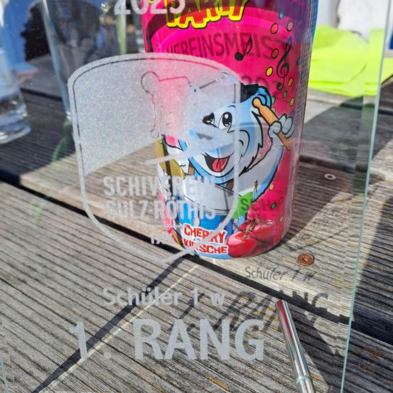 Eine Nahaufnahme eines gravierten Glases mit 'Schuler 1. Rang'. Dahinter ist ein rosa Getränk mit einem Cartoon-Charakter und Musiknoten zu sehen. Der Hintergrund zeigt eine verschwommene Ansicht eines Tisches und eines Glases.