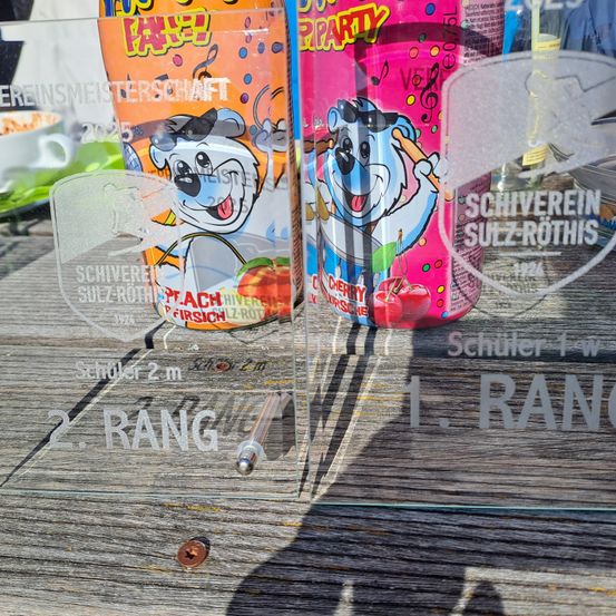 Ein Glasdisplay zeigt Auszeichnungen mit einem Hund, der einen Drumstick hält, beschriftet mit 'Schüler 2m' und '2. RANGE'. Daneben stehen zwei Getränkedosen, eine pfirsich- und eine kirschgeschmackige, beide mit der Marke 'SCHWEREIN SULZ-ROTHIS'. Der Hintergrund ist unscharf.