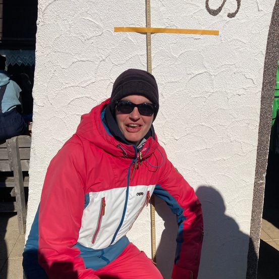 Ein Mann in einer roten und weißen Skijacke und Sonnenbrille posiert vor einer strukturierten weißen Wand mit einem gelben Maßband.