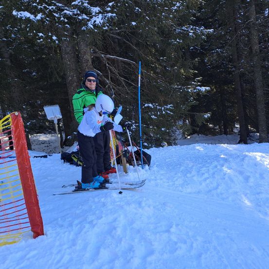 Eine Person mit weißem Helm ski fährt auf schneebedecktem Boden. Ein blauer Pfosten steht vor ihnen. Ein Mann in einer grünen Jacke steht hinter ihnen.