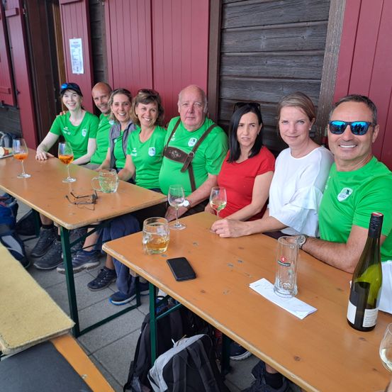 Eine Gruppe von Menschen in grünen T-Shirts sitzt an einem Tisch und lächelt. Weingläser, ein Handy und eine Flasche Wein stehen auf dem Tisch. Sie sind in einem Restaurant.