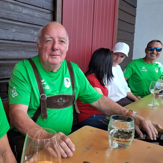 Ein älterer Mann in einem grünen T-Shirt und einer Lederhose sitzt an einem Tisch mit einem Glas Wasser und einem weiteren Glas mit einer Zitronenscheibe. Hinter ihm sitzen eine Frau in einem roten T-Shirt und ein weiterer Mann in einem grünen T-Shirt mit einer weißen Kappe. Eine Frau mit langen Haaren ist ebenfalls anwesend.