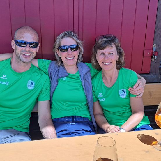 Drei Personen sitzen zusammen auf einer Bank draußen, tragen grüne T-Shirts mit Logos und lächeln für ein Foto. Sie sind in der Nähe einer roten Tür mit einem Schloss. Auf dem Tisch stehen Getränke in Gläsern.