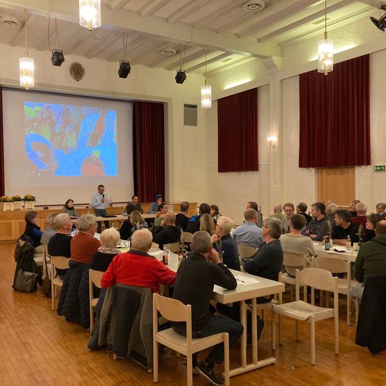 Ein Raum mit einer Projektionsleinwand zeigt ein Bild, während ein Mann spricht. Viele Menschen sitzen an Tischen vor der Leinwand.