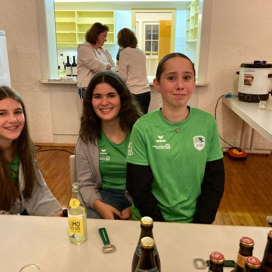 Drei junge Frauen in grünen T-Shirts sitzen an einem Tisch mit Getränken vor ihnen. Eine hat einen Flaschenöffner. Hinter ihnen stehen zwei Frauen an einem Tresen, und eine Kaffeemaschine steht auf einem nahen Tisch.