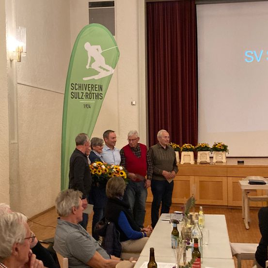 Eine Gruppe von Menschen steht in einem Raum mit einem Banner hinter ihnen. Sie stehen vor einer Bühne mit einer Projektionsleinwand. Ein Tisch mit Blumen und Flaschen steht rechts.
