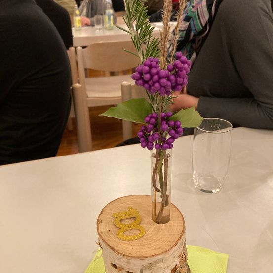 Ein Tischdekoration besteht aus einer hölzernen Log-Basis mit der Nummer '8', die eine klare Vase mit einem lila Blumenarrangement hält. In der Nähe befindet sich ein klares Glas und eine verschwommene Ansicht von sitzenden Personen.