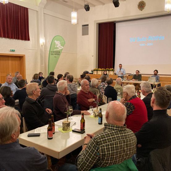 Ein Raum mit einer Projektionsleinwand zeigt SV Sulz-Rothis 1925-2025. Ein Mann spricht an einem Podium mit zwei anderen. Ein langer Tisch mit Wein- und Bierflaschen steht vor einem sitzenden Publikum.