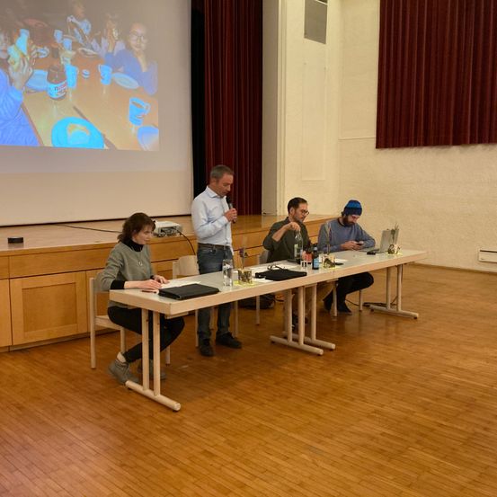 Eine Gruppe von Menschen sitzt an einem langen Tisch, wobei einer der Männer ein Mikrofon hält. Sie sehen einem Projektorbildschirm zu, der ein Bild von Kindern zeigt.