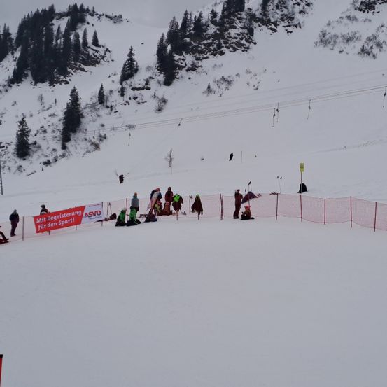 Eine Gruppe von Menschen versammelt sich auf einem verschneiten Berghang. Ein rotes Banner mit der Aufschrift 'Mit Registrierung für den Sport!' ist zu sehen. Mehrere Personen stehen und sitzen herum, wobei im Hintergrund Skilifte zu erkennen sind.