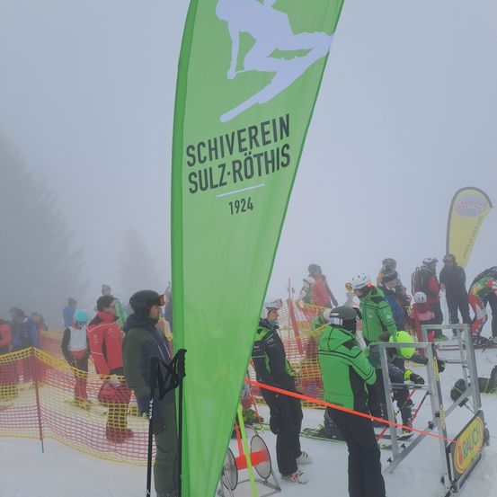 Eine Gruppe von Menschen versammelt sich in einem verschneiten Bereich. Sie tragen Winterkleidung und Helme. Ein grüner Banner mit dem Text SCHIVEREI SULZ-ROTHIS 1924 ist sichtbar. Einige Leute stehen in der Nähe eines Zauns, andere stehen in einer Reihe. Eine Person hält einen Skistock.