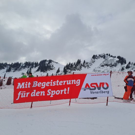 Ein Banner vor verschneiten Bergen trägt die Aufschrift 'Mit Begeisterung für den Sport!' in weißen Buchstaben auf rotem Hintergrund. Ein Mensch steht in der Nähe.