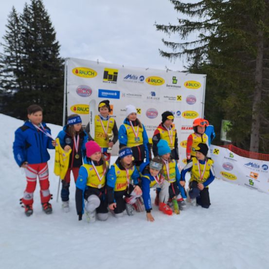 Eine Gruppe junger Athleten in gelben Westen und Skiausrüstung posiert für ein Foto auf einem verschneiten Hang mit einem Banner dahinter.
