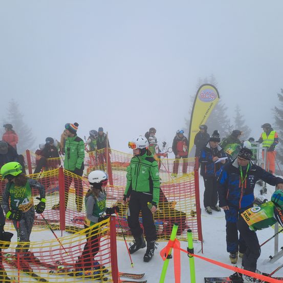 Skifahrer versammeln sich in einem nebligen Bereich, einige tragen grüne Uniformen mit Nummern. Sie halten Skistöcke und befinden sich in der Nähe eines gelb-roten Netzes. Dahinter stehen Menschen in einer Reihe, einige tragen reflektierende Westen. Ein gelber Banner mit Text ist sichtbar, und Bäume umgeben den Bereich.