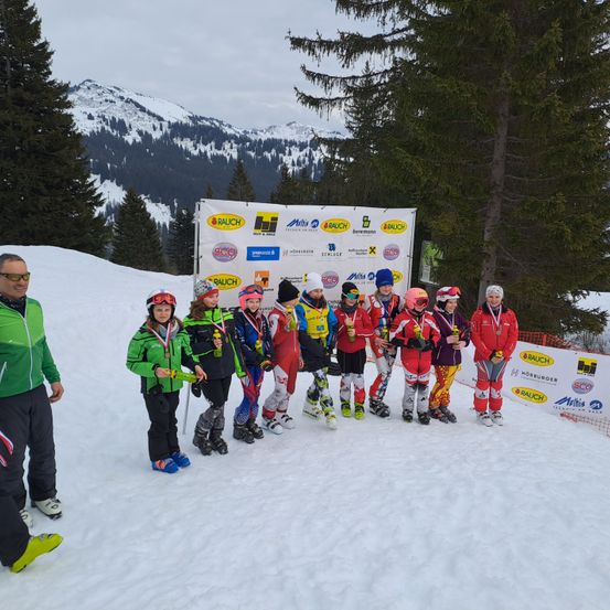 Eine Gruppe junger Skirennläufer in bunten Skianzügen steht in einer Reihe im Schnee mit Medaillen um den Hals. Sie stehen vor einem Banner.