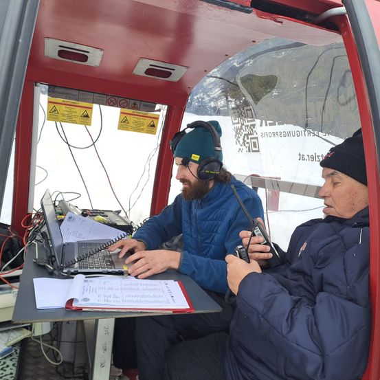 Zwei Männer in Winterkleidung sitzen in einer roten Kabine mit Glasfront und verwenden Funkgeräte. Ein Laptop und Papiere liegen auf dem Tisch vor ihnen.