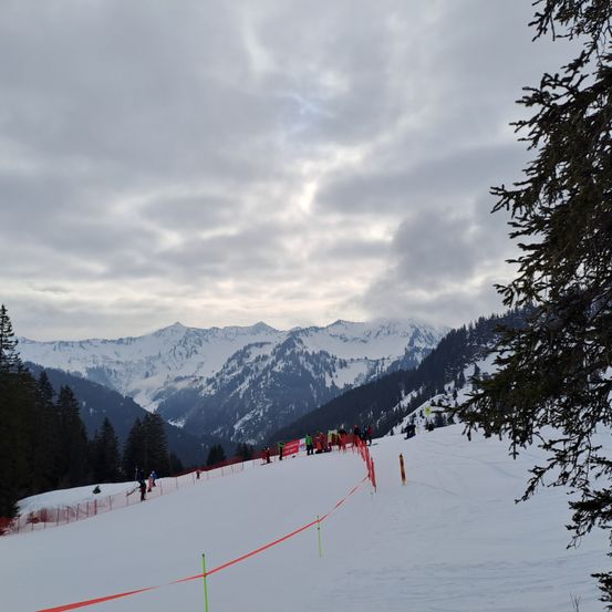 Verschneite Berge unter einem bewölkten Himmel mit einer Ski-Piste und einer Gruppe von Leuten in roten Jacken und Skiausrüstung.