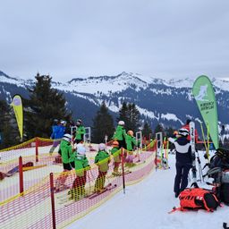 Eine Gruppe von Kindern in grünen Skijacken steht in einer Reihe auf einem verschneiten Berg. Sie halten Skistöcke und stehen in der Nähe eines gelben und roten Netzes. Im Hintergrund befinden sich grüne Banner und eine Person, die Skier hält.