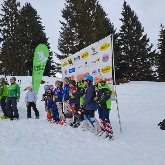 Eine Gruppe von Kindern in Winterkleidung steht in einer Reihe, möglicherweise nach einem Wettkampf, mit Schnee und immergrünen Bäumen im Hintergrund.
